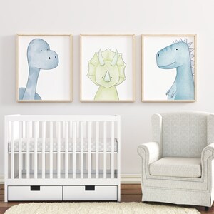May include: Three watercolor dinosaur prints in light blue, green, and yellow colors. The prints are framed in natural wood and hung on a white wall above a white crib and a white armchair.