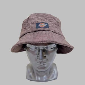 May include: A brown corduroy bucket hat with a small rectangular patch featuring the Dickies logo in red, yellow, and blue. The hat has a wide brim and a textured, ribbed fabric. The hat is displayed on a mannequin head.