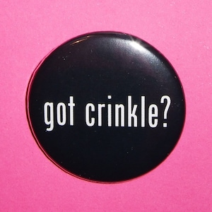 Puede incluir: Un botón negro con texto blanco que dice "got crinkle?" El botón está sobre un fondo rosa.