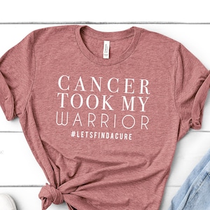 May include: A heather red t-shirt with the text "Cancer took my warrior #letsfindacure" printed in white. The shirt is laid flat on a white wooden surface with a pair of white sneakers, a pair of blue jeans, and three small potted succulents.