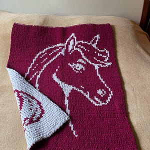 May include: A knitted burgundy blanket featuring a white horse head design. The blanket is folded, showing a grey section with a white heart. The blanket is on a tan surface.