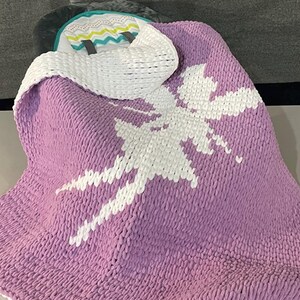 May include: A lavender knitted blanket with a large white fairy design. The blanket has a thick, textured appearance. A white knitted cowl with a patterned trim is draped over the blanket.
