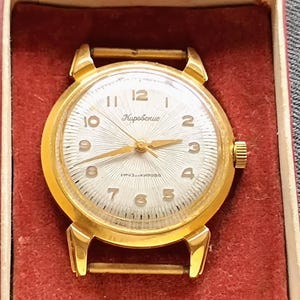 May include: A gold-toned wristwatch with a white face and gold hands and numerals. The watch face has a radial pattern and the text "Кировские" is visible. The watch is presented in a red box.