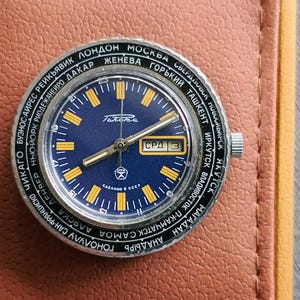 Puede incluir: Un reloj de pulsera plateado con una esfera azul y marcas amarillas. El reloj tiene un bisel giratorio con nombres de ciudades impresos alrededor del borde. La esfera del reloj tiene una ventana de fecha y una ventana de día de la semana. La esfera del reloj también tiene la palabra "СССР" impresa en ella.