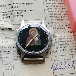 May include: A vintage wristwatch with a silver-toned case and a black face featuring an icon of a religious figure. The watch rests on a document with Cyrillic text and is accompanied by a pink rectangular object.