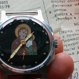 May include: A vintage wristwatch with a silver-tone case and a black face featuring an icon of a religious figure. The watch has gold-tone hands and hour markers. The watch is held in a hand, with a document in the background.