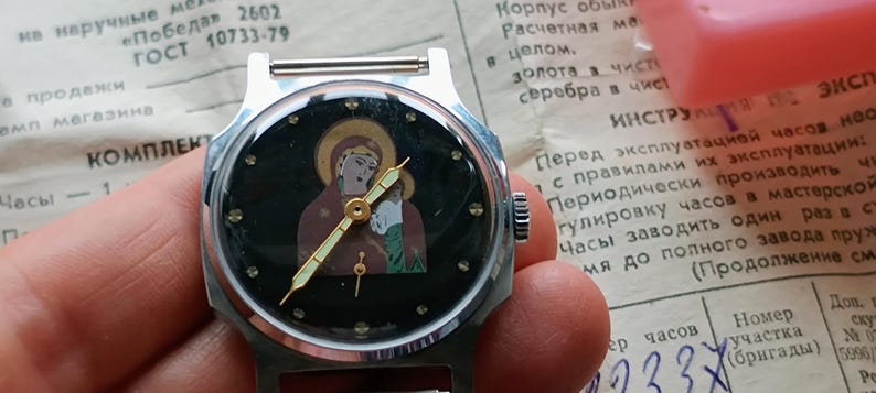 May include: A vintage wristwatch with a silver-tone case and a black face featuring an icon of a religious figure. The watch has gold-tone hands and hour markers. The watch is held in a hand, with a document in the background.