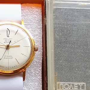 Può includere: Orologio da polso dorato vintage con cinturino bianco, presentato nella sua scatola originale. Il quadrante color crema presenta lancette e indici dorati, con la scritta "POLJOT de luxe automatic". L'orologio è in una custodia di plastica trasparente.