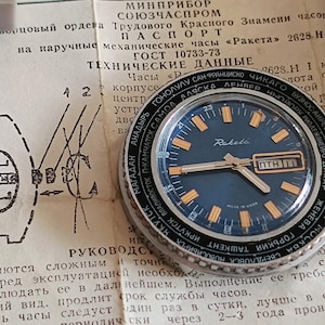 May include: A vintage Raketa wristwatch with a dark blue face, gold-coloured hour markers, and a day-date display. The watch has a black bezel with city names and is set against old documents.