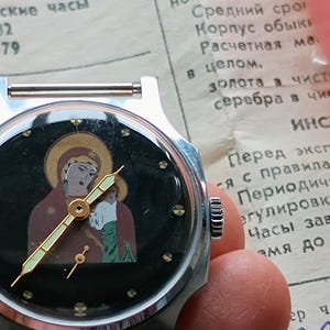 May include: A vintage silver-toned wristwatch with a black face featuring an icon of the Virgin Mary and child. The watch has gold-colored hands and hour markers. The watch is held over a document with Russian text.