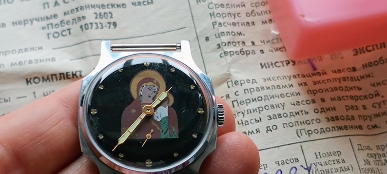 May include: A vintage silver-toned wristwatch with a black face featuring an icon of the Virgin Mary and child. The watch has gold-colored hands and hour markers. The watch is held over a document with Russian text.