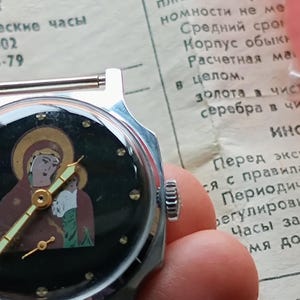 May include: A vintage silver wristwatch with a black face featuring an icon of a religious figure. The watch has gold-colored hands and hour markers. The watch is held in a hand, with a document in the background.