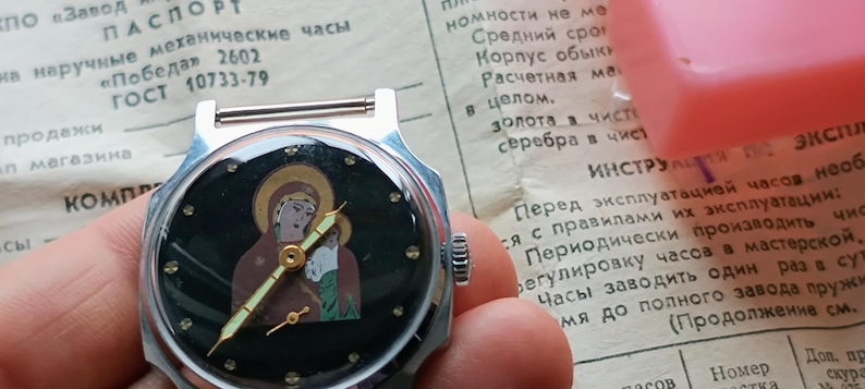 May include: A vintage silver wristwatch with a black face featuring an icon of a religious figure. The watch has gold-colored hands and hour markers. The watch is held in a hand, with a document in the background.