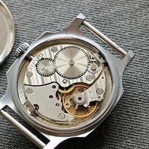 May include: A close-up view of the inner workings of a silver-toned wristwatch. The watch's back is open, revealing intricate gears, springs, and other mechanical components. The watch face is missing, and the back cover is placed to the side.