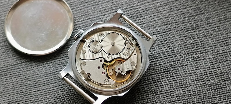 May include: A close-up view of the inner workings of a silver-toned wristwatch. The watch's back is open, revealing intricate gears, springs, and other mechanical components. The watch face is missing, and the back cover is placed to the side.
