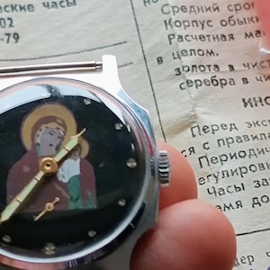 May include: A vintage wristwatch with a black face featuring a religious icon. The watch has gold-colored hands and a silver-colored case. The watch is displayed on a piece of paper with Russian text.