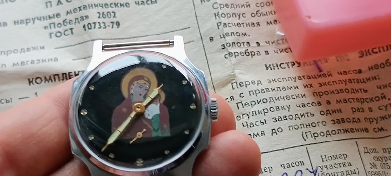 May include: A vintage wristwatch with a black face featuring a religious icon. The watch has gold-colored hands and a silver-colored case. The watch is displayed on a piece of paper with Russian text.