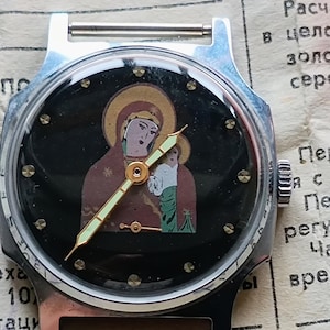 May include: A vintage wristwatch with a black face featuring an icon of the Virgin Mary and child. The watch has a silver-colored case and a black band. The hands are gold-colored.