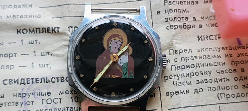 May include: A vintage wristwatch with a black face featuring an icon of the Virgin Mary and child. The watch has a silver-colored case and a black band. The hands are gold-colored.