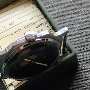 May include: A vintage wristwatch with a black face and silver-toned casing rests inside a green box. The watch has a black band and is partially obscuring a piece of paper with text. The watch is a classic timepiece.