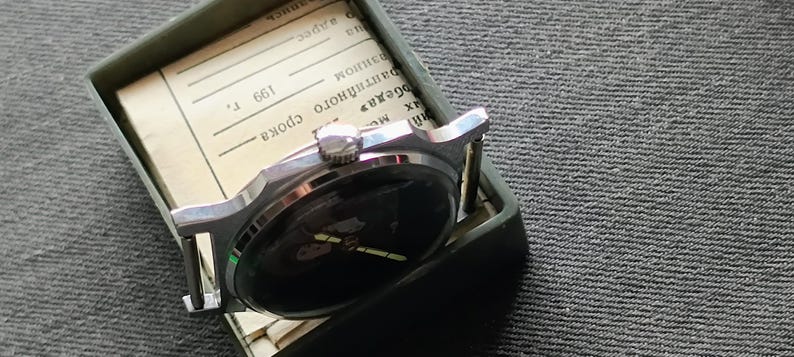 May include: A vintage wristwatch with a black face and silver-toned casing rests inside a green box. The watch has a black band and is partially obscuring a piece of paper with text. The watch is a classic timepiece.