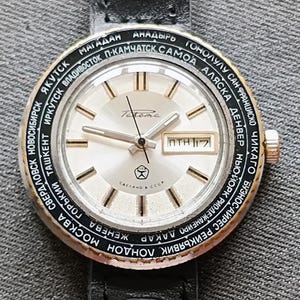 May include: A vintage silver watch with a white face, the brand name "Tastema" and a date display. The watch has a black bezel with city names and a black leather strap. The watch face has gold-toned hour markers and hands.