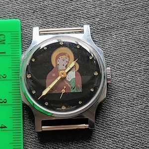 May include: A silver-toned wristwatch with a black face featuring a religious icon. The watch has a round face with a silver bezel and a black band. A green ruler with the text "COOLFORSC" is next to the watch.