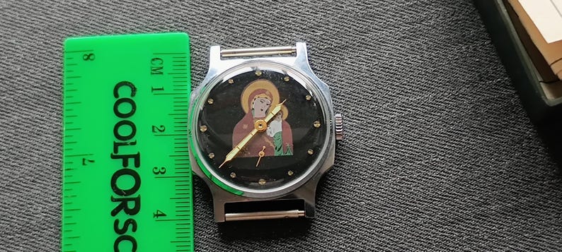 May include: A silver-toned wristwatch with a black face featuring a religious icon. The watch has a round face with a silver bezel and a black band. A green ruler with the text "COOLFORSC" is next to the watch.