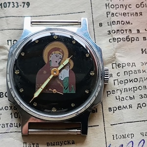 May include: A vintage wristwatch with a black face featuring an image of a religious figure. The watch has a silver-tone case and a brown strap. The watch is set on a piece of paper with Russian text.