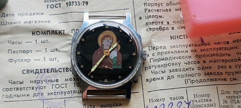 May include: A vintage wristwatch with a black face featuring an image of a religious figure. The watch has a silver-tone case and a brown strap. The watch is set on a piece of paper with Russian text.