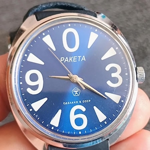 Peut inclure: Une montre-bracelet Raketa avec un boîtier rond argenté et un cadran bleu foncé. Le cadran présente de grands chiffres arabes blancs aux positions 0, 3, 6 et 9, des index triangulaires blancs, des aiguilles dorées et le texte Raketa et СДЕЛАНО В СССР.