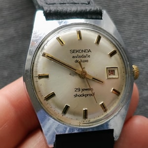 Può includere: Un orologio Sekonda argentato con quadrante bianco e dettagli dorati. L'orologio ha una finestrella per la data e riporta la scritta "aulodate de-luxe" e "29 jewels shockproof".