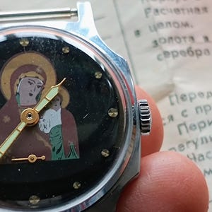 May include: A vintage wristwatch with a black face featuring an icon of a religious figure. The watch has a silver-colored metal case and a gold-colored hour and minute hands. The watch is held in a hand.