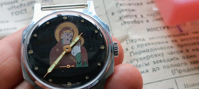May include: A vintage wristwatch with a black face featuring an icon of a religious figure. The watch has a silver-colored metal case and a gold-colored hour and minute hands. The watch is held in a hand.