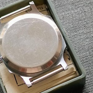 May include: A silver-toned watch case with a circular back, resting inside a green rectangular box. The watch case has two silver bars for the watch band. The box contains a folded paper with printed text. The background is a gray textured fabric.