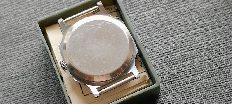 May include: A silver-toned watch case with a circular back, resting inside a green rectangular box. The watch case has two silver bars for the watch band. The box contains a folded paper with printed text. The background is a gray textured fabric.