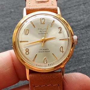 Può includere: Un orologio da polso vintage color oro con quadrante chiaro e cinturino in pelle marrone. Il quadrante dell'orologio presenta il testo "POLJOT de luxe automatic" e "29 jewels shock-resist". L'orologio ha lancette e indici dorati.