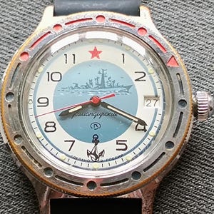 Puede incluir: Un reloj de pulsera vintage con una esfera azul claro que presenta una ilustración de un barco y la palabra "Командирские". El reloj tiene una estrella roja a las 12, una ventana de fecha y un bisel plateado con detalles en rojo. La correa es gris oscuro.