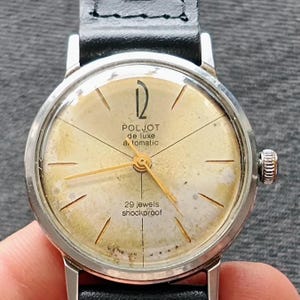Puede incluir: Reloj de pulsera automático Poljot de Luxe vintage con caja plateada y esfera dorada. El reloj presenta agujas e indicadores dorados, una correa de cuero negro y el texto "29 jewels shockproof".