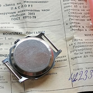 May include: A vintage wristwatch with a silver-colored back casing, lying on a document with Cyrillic text. The watch is next to a small, dark green box and a pink plastic container. The document includes the text "Pobeda 2602".