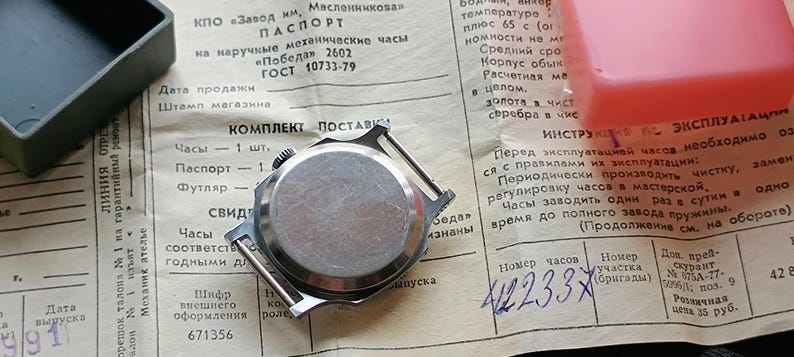 May include: A vintage wristwatch with a silver-colored back casing, lying on a document with Cyrillic text. The watch is next to a small, dark green box and a pink plastic container. The document includes the text "Pobeda 2602".