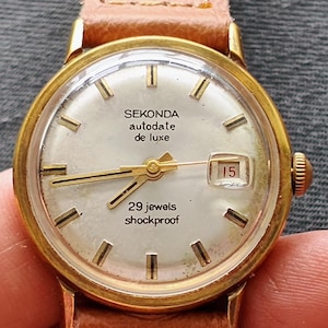 Può includere: Un orologio da polso vintage Sekonda autode date de luxe con cassa color oro e quadrante chiaro. L'orologio presenta lancette e indici dorati, un datario che mostra il numero 15 e un cinturino in pelle marrone. Il quadrante recita "29 jewels shockproof".