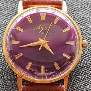 May include: Vintage wristwatch with a deep purple face, gold hands, and gold hour markers. The watch has a gold-tone case and a brown leather band. The face has the word "Луч" in gold script. The watch is a classic timepiece.