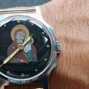 May include: A silver wristwatch with a black face featuring an icon of a religious figure. The watch has gold hands and a silver band. The watch face is surrounded by small, clear crystals.
