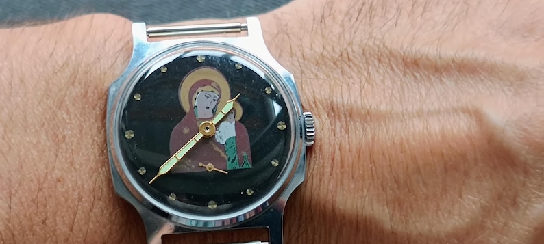 May include: A silver wristwatch with a black face featuring an icon of a religious figure. The watch has gold hands and a silver band. The watch face is surrounded by small, clear crystals.