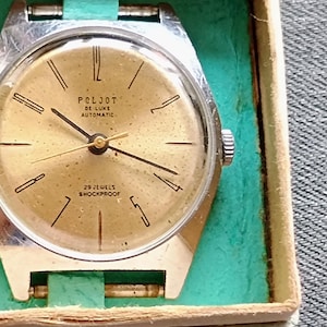 Può includere: Un orologio vintage color oro con cassa argentata, presentato nella sua scatola originale. Il quadrante dell'orologio mostra il marchio "Poljot" e le parole "De Luxe Automatic" e "29 Jewels Shockproof". L'orologio è posto su uno sfondo turchese.