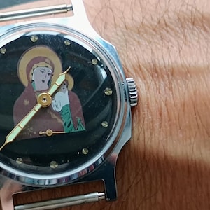 May include: A silver-toned wristwatch with a black face featuring a religious icon of a woman and child. The watch has a round face with gold-colored hands and hour markers. The watch is on a silver band.