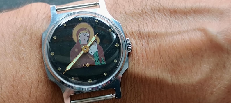 May include: A silver-toned wristwatch with a black face featuring a religious icon of a woman and child. The watch has a round face with gold-colored hands and hour markers. The watch is on a silver band.