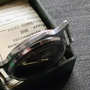 May include: A silver-toned wristwatch with a black face rests inside a green rectangular box. The watch is partially visible, showing its face and band. The box contains a stack of paper with text. The background is a textured gray surface.