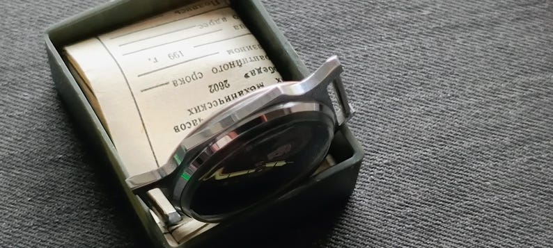 May include: A silver-toned wristwatch with a black face rests inside a green rectangular box. The watch is partially visible, showing its face and band. The box contains a stack of paper with text. The background is a textured gray surface.